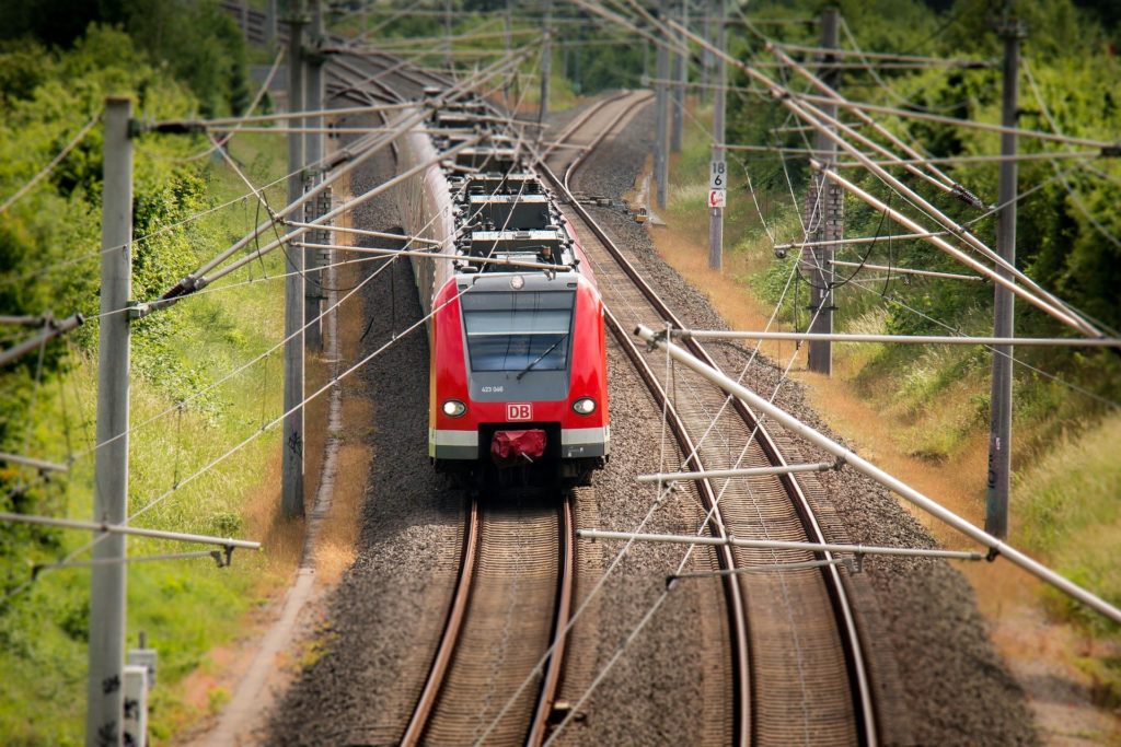 Roter, moderner Regionalzug fährt auf einer elektrifizierter Bahnstrecke auf freier, bebuschter Strecke. 