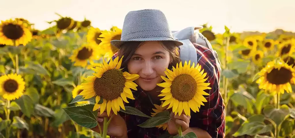 Junge Frau im rot/blau karriertem Hemd - mit viel Lust auf Ortsverbandsgründung - steht gebeugt mitten in einem Sonnenblumenfeld und possiert mit den Sonnenblumenköpfen. Auf dem Kop trägt sie einen grau karrierten Hut. Auf dem Rücken trägt sie einen blaugrauen Rucksack.