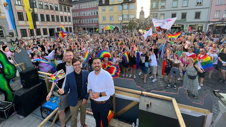 3 Personen stehen vor einer großen Menge Menschen auf einer Bühne und schauen in die Kamera. Einige schwenken die Regenbogen-Fahnen der Queer-Community.